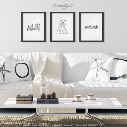 A group of three framed drawings on a white wall hanging above a couch with pillows and a blanket. The line art drawings include Harness Racing, a Persian cat, and the Portland Skyline