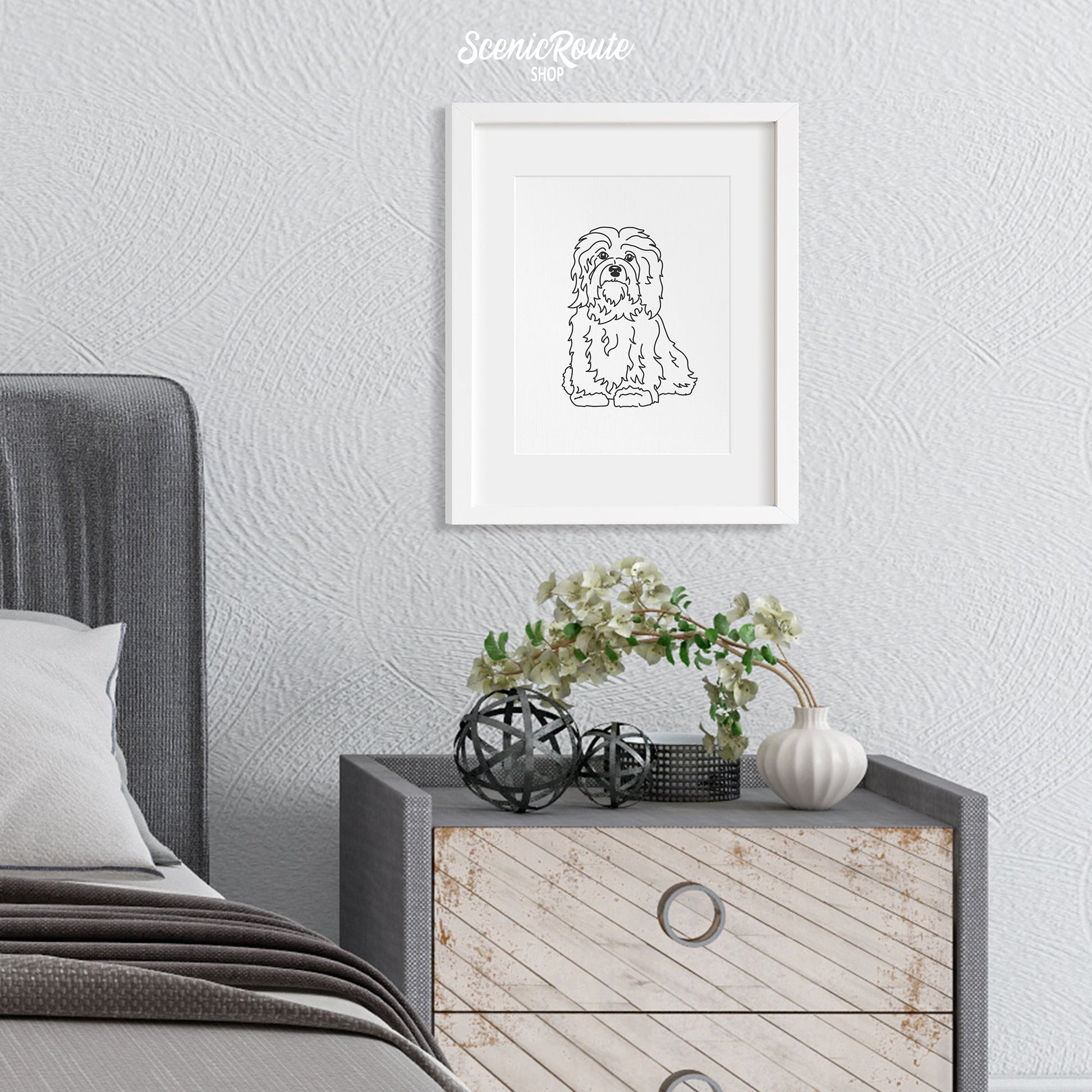 A framed line art drawing of a Havanese dog above a nightstand next to a bed