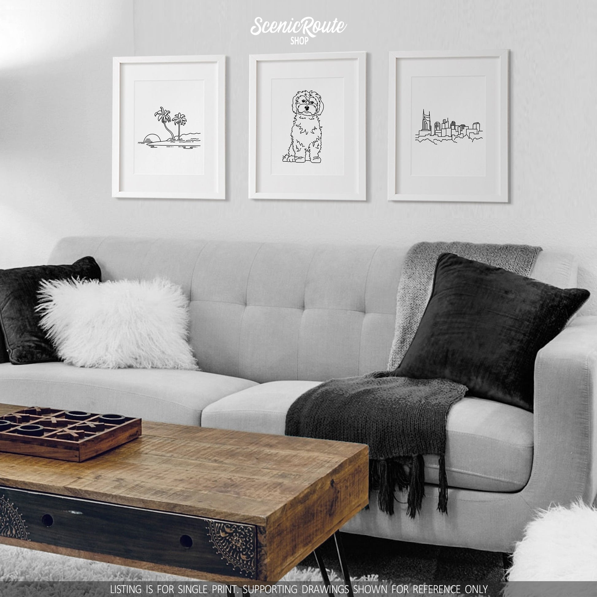 A group of three framed drawings on a white wall hanging above a couch with pillows and a blanket. The line art drawings include an Island, a Maltese dog, and the Nashville Skyline