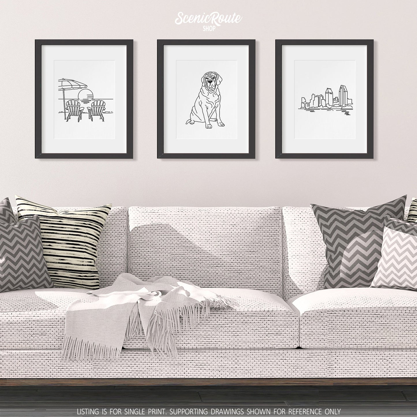 A group of three framed drawings on a white wall hanging above a couch with pillows and a blanket. The line art drawings include Adirondack Beach Chairs, a Mastiff dog, and the San Diego Skyline