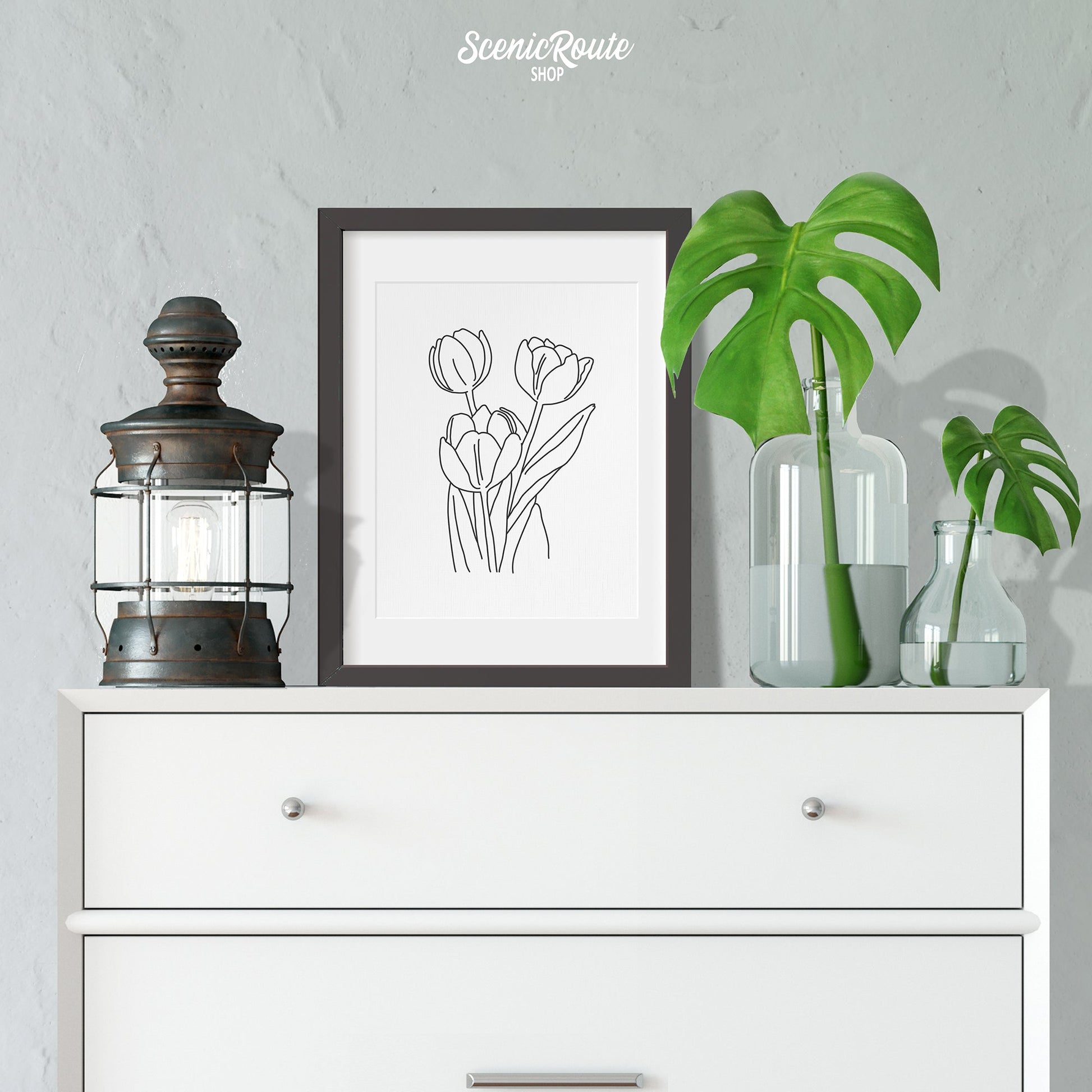 A framed line art drawing of a Tulip Flower on a white dresser with plants and a lantern