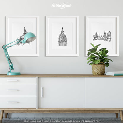 A group of three framed drawings on a wall hanging above a credenza with a lamp and a potted plant. The line art drawings include the Eiffel Tower, Big Ben, and the London Skyline