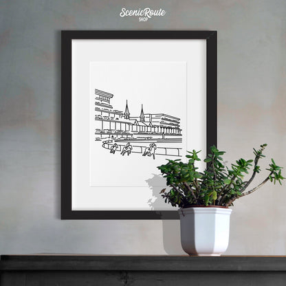 A framed line art drawing of Churchill Downs over a table with a plant