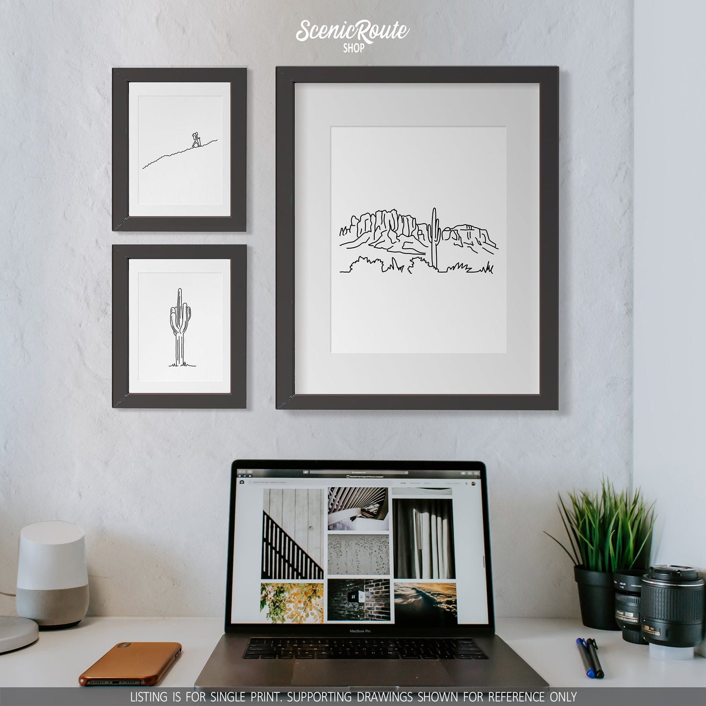 A group of three framed drawings on a wall above a desk and a laptop. The line art drawings include a person Hiking, a Saguaro Cactus, and the Superstition Mountains